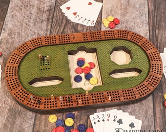 Könnte beinhalten: Ein hölzernes Cribbage-Brett mit einer grünen Spielfläche und einem braunen Holzrahmen. Das Brett hat eine eingebaute Punkteleiste, Stifte und ein Fach für Karten. Es gibt auch Spielkarten und Chips, die um das Brett herum verstreut sind.