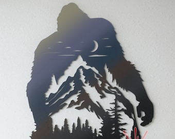 May include: Black metal silhouette of Bigfoot standing in front of a mountain range with a crescent moon in the sky. The silhouette is cut out of metal and has a rustic, weathered look. The text "TIMBERLAND CNC" is visible at the bottom of the image.