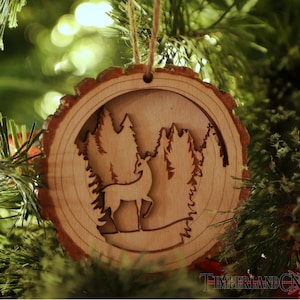 May include: A wooden Christmas ornament shaped like a tree trunk with a cutout design of a deer standing in a forest. The ornament is hanging on a Christmas tree.