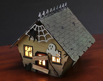 May include: A wooden Halloween house with a green roof, lit windows, and spooky decorations such as a spider web, a ghost, and a black cat. The house is sitting on a wooden surface.