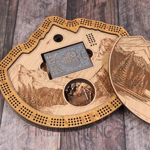 Può includere: Un tabellone da cribbage in legno con un design a tema montagna. Il tabellone è aperto e il coperchio è rimosso, rivelando un mazzo di carte e un vano per i birilli. Il tabellone è realizzato in legno chiaro e ha una tinta marrone scuro.