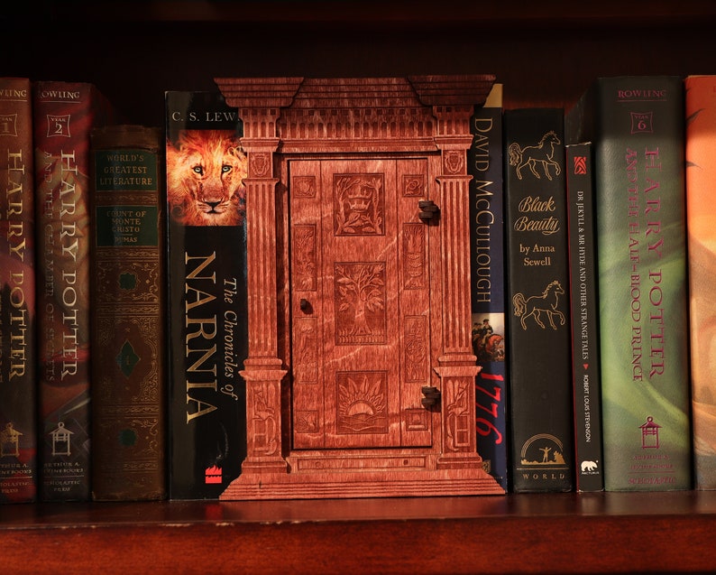 K&ouml;nnte beinhalten: Ein B&uuml;cherregal mit einer Auswahl an B&uuml;chern, darunter Harry Potter, Die Chroniken von Narnia und Black Beauty. Das B&uuml;cherregal ist aus dunklem Holz gefertigt und hat eine dekorative Holzt&uuml;r in der Mitte.