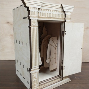 K&ouml;nnte beinhalten: Ein Holzmodell eines Kleiderschranks mit offener T&uuml;r, in dem ein kleiner Baum und zwei M&auml;ntel h&auml;ngen. Der Kleiderschrank hat ein detailliertes, verziertes Design mit S&auml;ulen und einem dekorativen Oberteil.