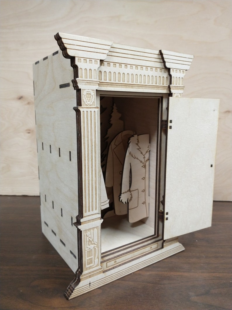 K&ouml;nnte beinhalten: Ein Holzmodell eines Kleiderschranks mit offener T&uuml;r, in dem ein kleiner Baum und zwei M&auml;ntel h&auml;ngen. Der Kleiderschrank hat ein detailliertes, verziertes Design mit S&auml;ulen und einem dekorativen Oberteil.