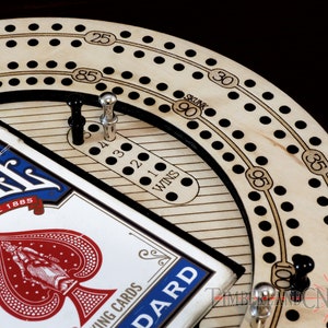 Puede incluir: Un tablero de cribbage de madera con puntos negros y blancos, una clavija negra y plateada y una baraja de cartas. El tablero tiene una pista circular con n&uacute;meros del 20 al 100, y una pista m&aacute;s peque&ntilde;a con n&uacute;meros del 1 al 4. El tablero tambi&eacute;n tiene una etiqueta "SKUNK" y "WINS".