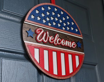Könnte beinhalten: Ein rundes, hölzernes Willkommensschild mit einem rot-weiß-blauen amerikanischen Flaggenmuster. Das Schild trägt den Schriftzug "Willkommen" in weißen Buchstaben auf braunem Hintergrund.