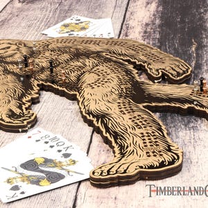 May include: A wooden game board shaped like a Bigfoot, with a brown and black design. The board has a grid of holes and is designed for a game of peg solitaire. The board is on a wooden surface with playing cards and game pieces.