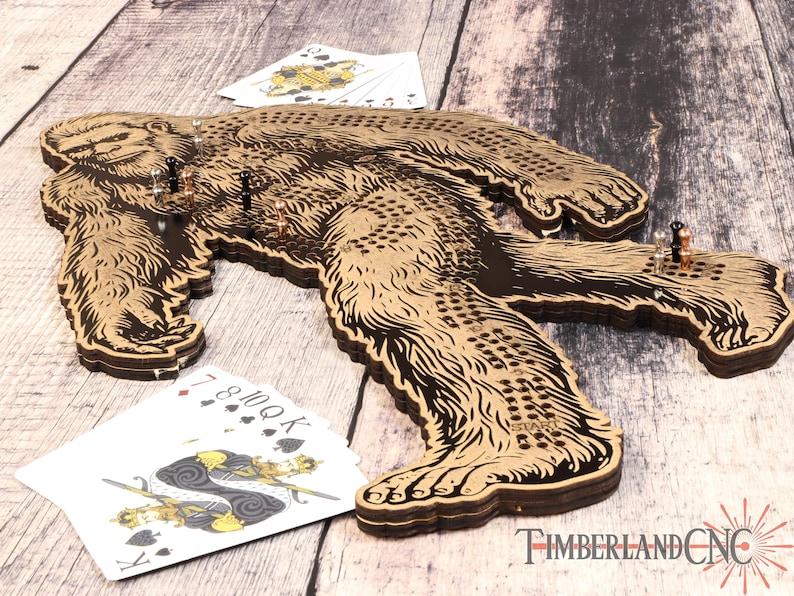 May include: A wooden game board shaped like a Bigfoot, with a brown and black design. The board has a grid of holes and is designed for a game of peg solitaire. The board is on a wooden surface with playing cards and game pieces.