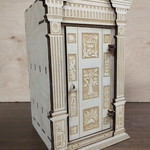 K&ouml;nnte beinhalten: Eine Holzkiste mit einer geschnitzten T&uuml;r mit aufwendigen Designs. Die Kiste ist in einer hellen Holzfarbe lackiert und hat einen dekorativen Rahmen oben.