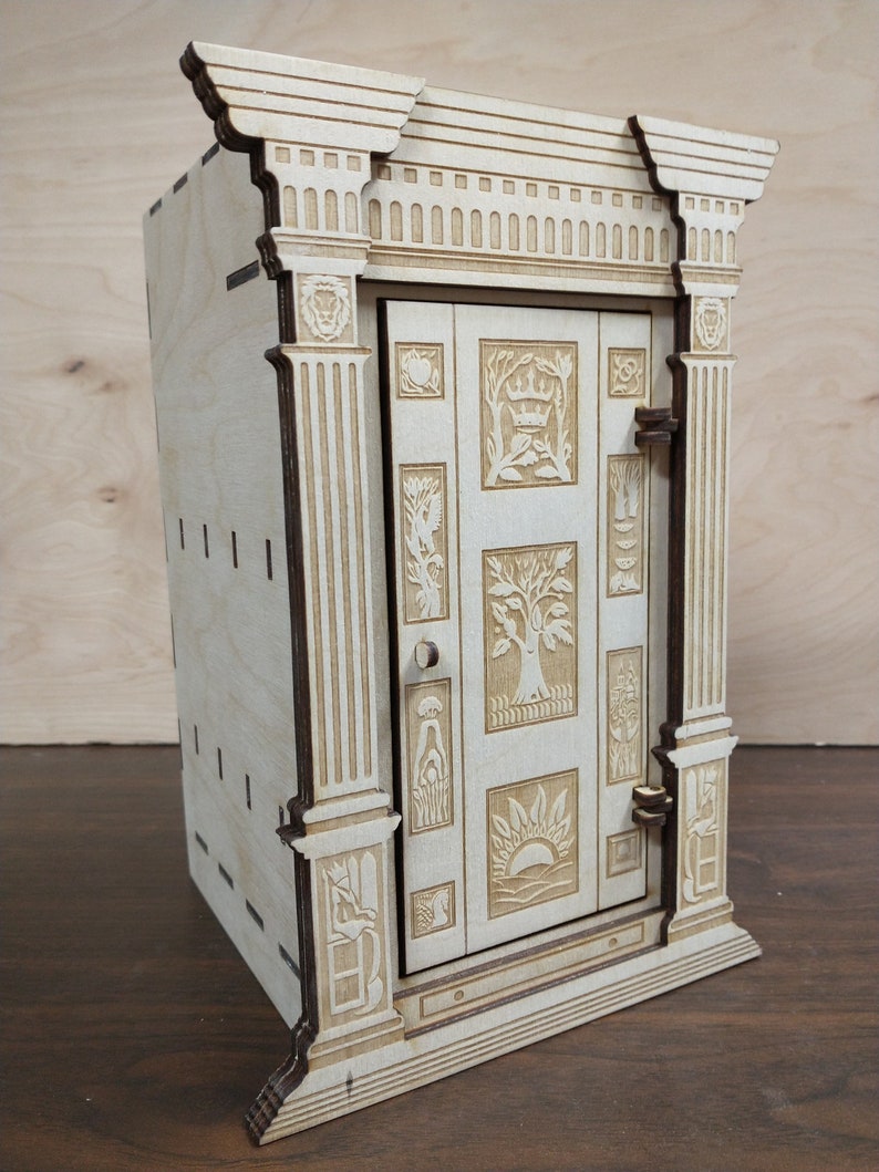 K&ouml;nnte beinhalten: Eine Holzkiste mit einer geschnitzten T&uuml;r mit aufwendigen Designs. Die Kiste ist in einer hellen Holzfarbe lackiert und hat einen dekorativen Rahmen oben.
