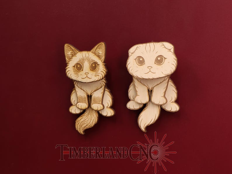 May include: Two light-colored wooden cat cutouts with engraved details. The left cat has pointed ears and a fluffy tail, while the right cat has folded ears and a smooth tail. The text TIMBERLANDCNC is visible at the bottom in red.