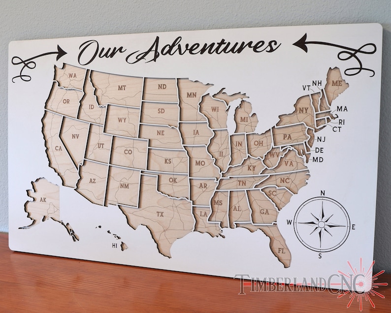 May include: A white wooden map of the United States with each state outlined and labeled. The map is mounted on a white background with the words "Our Adventures" written in cursive above the map. A compass is in the lower right corner of the map.