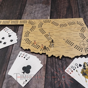 May include: A wooden game board shaped like the state of Oklahoma with a compass in the bottom right corner. The board has holes for pegs and is labeled with cities and towns in Oklahoma. There are playing cards on the table next to the board.