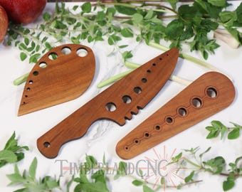 May include: Three wooden kitchen utensils with cutouts. The utensils are made of light brown wood and have a variety of shapes and sizes. The utensils are arranged on a white surface with green leafy plants.
