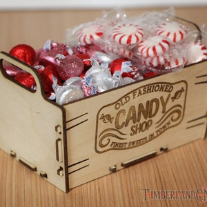 Puede incluir: Una caja de madera con un asa recortada, llena de chocolates envueltos en papel de aluminio rojo y rosa y caramelos de menta blanca y roja. La caja está grabada con el texto "Old Fashioned Candy Shop Finest Sweets in Town".