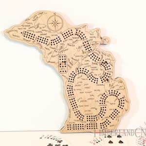 Op de afbeelding: Een houten spelbord in de vorm van de staat Michigan met de namen van steden en dorpen erop gedrukt. Het bord heeft een raster van gaten om een spel te spelen dat lijkt op een traditioneel damspel. Het bord is gemaakt van lichtbruin hout en heeft een natuurlijke afwerking. Het bord is omgeven door een witte achtergrond.