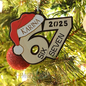 May include: A wooden Christmas tree ornament featuring a red Santa hat and a red glitter ball. The ornament is in the shape of the number 67, with the name "KARINA" and the year "2025" printed on it. The words "SIX" and "SEVEN" are also printed.