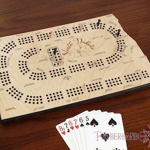 May include: A wooden game board shaped like the state of North Dakota. The board has a game track with holes for pegs and the names of cities in North Dakota printed on it. The board is made of light wood with a dark wood border. There are playing cards on the table next to the board.