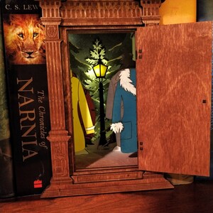 K&ouml;nnte beinhalten: Ein h&ouml;lzerner Kleiderschrank-Buchst&uuml;tze mit einer T&uuml;r, die sich &ouml;ffnet und eine magische Szene enth&uuml;llt. Die Szene beinhaltet einen Laternenpfahl, einen Baum und zwei Figuren, die M&auml;ntel tragen. Der Buchst&uuml;tze steht auf einem B&uuml;cherregal mit anderen B&uuml;chern.