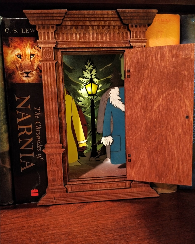 K&ouml;nnte beinhalten: Ein h&ouml;lzerner Kleiderschrank-Buchst&uuml;tze mit einer T&uuml;r, die sich &ouml;ffnet und eine magische Szene enth&uuml;llt. Die Szene beinhaltet einen Laternenpfahl, einen Baum und zwei Figuren, die M&auml;ntel tragen. Der Buchst&uuml;tze steht auf einem B&uuml;cherregal mit anderen B&uuml;chern.