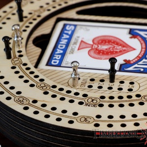 Puede incluir: Un tablero de cribbage de madera con una pista circular y agujeros numerados. El tablero est&aacute; hecho de madera clara con n&uacute;meros y puntos negros. Una carta de juego es visible en el centro del tablero, mostrando el As de Corazones de una baraja de cartas Bicycle.