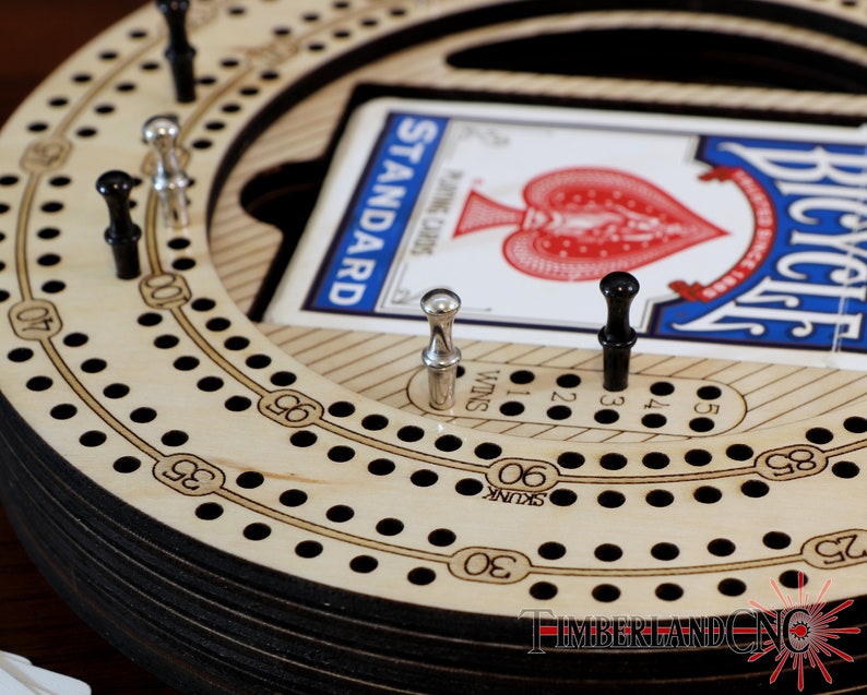 Puede incluir: Un tablero de cribbage de madera con una pista circular y agujeros numerados. El tablero est&aacute; hecho de madera clara con n&uacute;meros y puntos negros. Una carta de juego es visible en el centro del tablero, mostrando el As de Corazones de una baraja de cartas Bicycle.