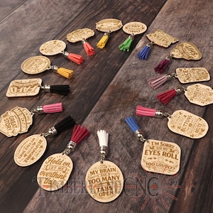 May include: A collection of wooden keychains with various engraved phrases and colorful tassels. The keychains have different shapes, including oval, rectangular, and irregular, and feature phrases like "Hold on, let me overthink this" and "I'm sorry did my eyes roll too loudly?"
