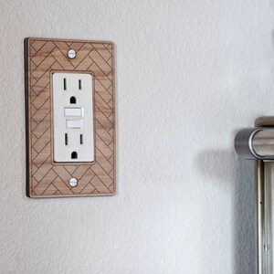 May include: A wooden outlet cover with a light brown wood grain finish and a geometric pattern. The outlet cover is designed to fit a standard US electrical outlet.