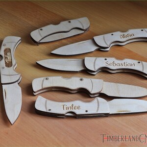 May include: A collection of wooden pocket knives, some engraved with names like "Alivia", "Sebastian", and "Tinlee". The knives are light brown and feature a classic folding design. The image is taken on a wooden surface.
