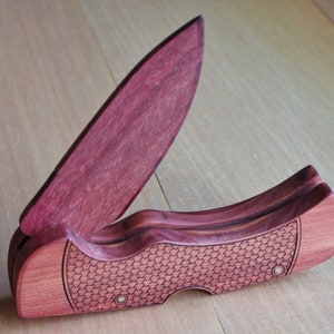 May include: A wooden folding knife with a partially open, reddish-purple blade. The handle is a lighter reddish-brown with a textured, honeycomb-like pattern. The knife is set against a light wood grain background.