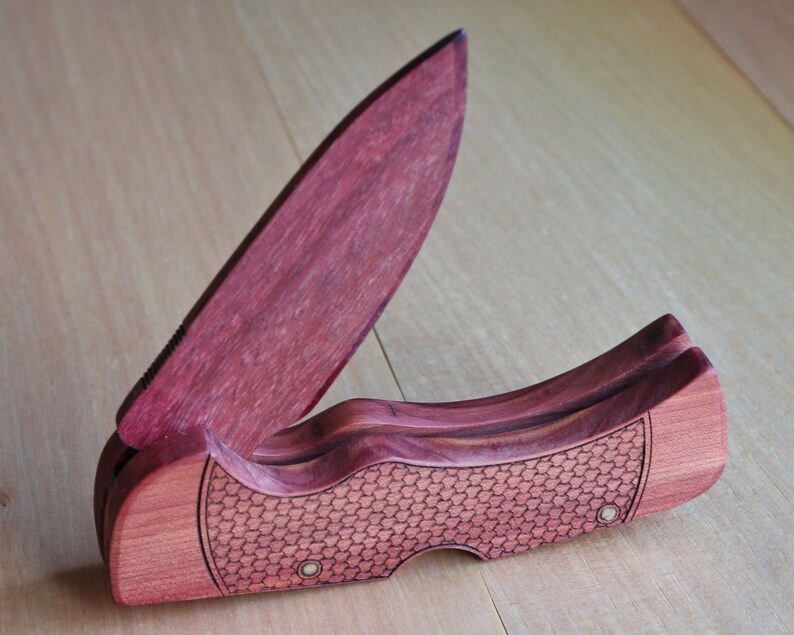 May include: A wooden folding knife with a partially open, reddish-purple blade. The handle is a lighter reddish-brown with a textured, honeycomb-like pattern. The knife is set against a light wood grain background.
