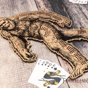 May include: A wooden Bigfoot-shaped game board with a black and brown design. The board has a series of holes for playing a game similar to a peg game. The board is on a wooden surface with playing cards and game pieces.