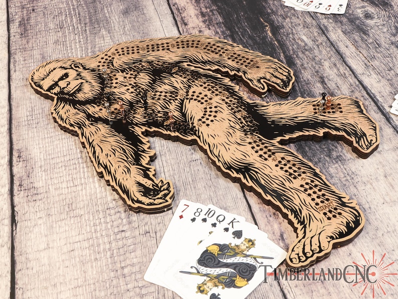 May include: A wooden Bigfoot-shaped game board with a black and brown design. The board has a series of holes for playing a game similar to a peg game. The board is on a wooden surface with playing cards and game pieces.