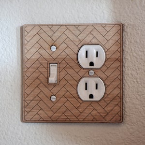 May include: A wooden wall plate with a herringbone pattern, featuring a light switch and a double electrical outlet.