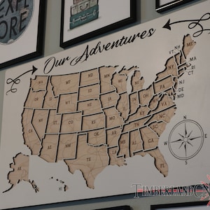 May include: A wooden map of the United States with the text "Our Adventures" at the top. Each state is outlined and labeled with its abbreviation. The map includes Alaska and Hawaii. A compass is in the lower right corner.