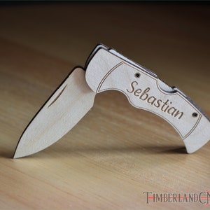 May include: A wooden folding knife with the name "Sebastian" engraved on the handle. The knife is made of light-colored wood and has a silver-colored blade. The knife is open, displaying the blade. The Timberland CNC logo is visible.