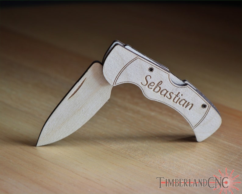 May include: A wooden folding knife with the name "Sebastian" engraved on the handle. The knife is made of light-colored wood and has a silver-colored blade. The knife is open, displaying the blade. The Timberland CNC logo is visible.