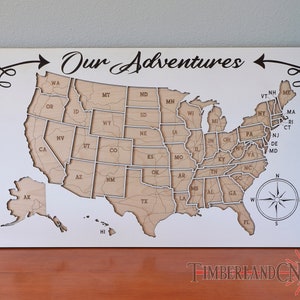 May include: A white wooden map of the United States with the text "Our Adventures" at the top. The map is divided into individual states, each with its abbreviation. The map is surrounded by a decorative border.