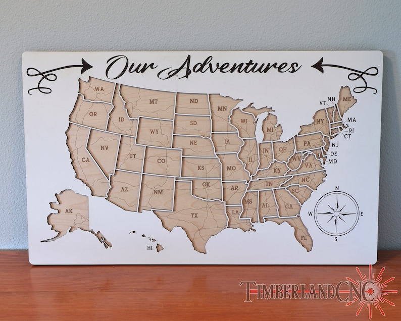 May include: A white wooden map of the United States with the text "Our Adventures" at the top. The map is divided into individual states, each with its abbreviation. The map is surrounded by a decorative border.