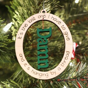 May include: A wooden Christmas ornament with the text "It's the last one I have to give and it's hanging by a thread" and the word "Damn" in green inside the circle.