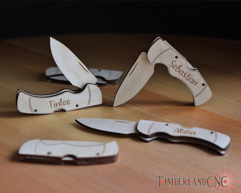 May include: A collection of personalized wooden pocket knives, some with blades open, others closed. The names "Tinlee", "Sebastian", and "Alivia" are engraved on the handles. The knives are light brown with dark brown accents.