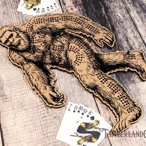 May include: A wooden Bigfoot game board with a brown and black illustration of Bigfoot walking. The board has a grid of holes for playing a game with marbles or pegs. The board is surrounded by playing cards.