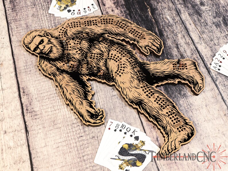 May include: A wooden Bigfoot game board with a brown and black illustration of Bigfoot walking. The board has a grid of holes for playing a game with marbles or pegs. The board is surrounded by playing cards.