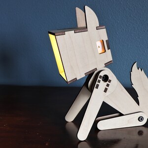 以下が含まれることがあります： 頭から光が漏れている木製の犬のランプ。犬は木製のテーブルの上に座っています。