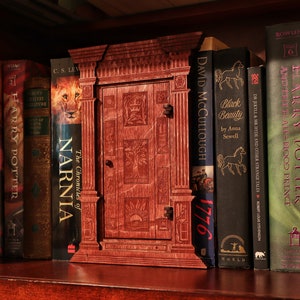 K&ouml;nnte beinhalten: Ein h&ouml;lzerner Buchst&uuml;tze in Form einer T&uuml;r mit einem geschnitzten Design. Der Buchst&uuml;tze steht in einem B&uuml;cherregal mit mehreren B&uuml;chern, darunter "Die Chroniken von Narnia", "Harry Potter" und "Black Beauty".