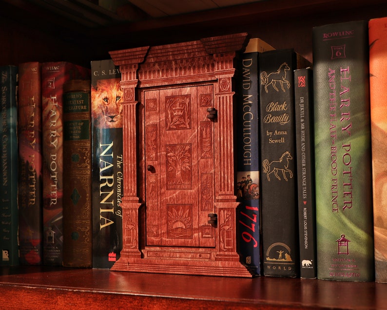 K&ouml;nnte beinhalten: Ein h&ouml;lzerner Buchst&uuml;tze in Form einer T&uuml;r mit einem geschnitzten Design. Der Buchst&uuml;tze steht in einem B&uuml;cherregal mit mehreren B&uuml;chern, darunter "Die Chroniken von Narnia", "Harry Potter" und "Black Beauty".