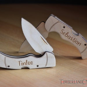 May include: Two wooden pocket knives, one open and one closed, with the names "Tinlee" and "Sebastian" engraved on them. The knives are made of light-colored wood and are set against a blurred background.