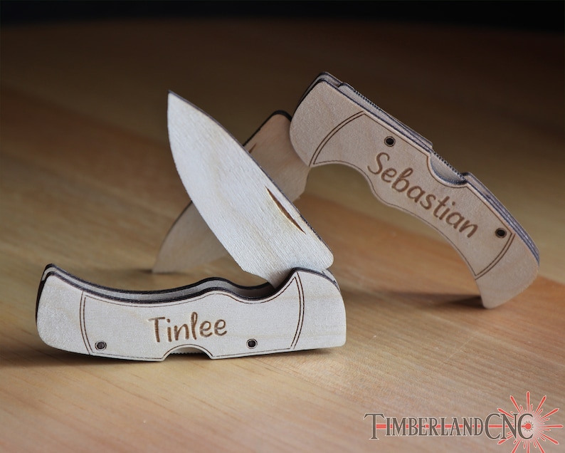 May include: Two wooden pocket knives, one open and one closed, with the names "Tinlee" and "Sebastian" engraved on them. The knives are made of light-colored wood and are set against a blurred background.