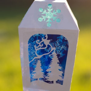 May include: White paper lantern with a blue and white snowflake design on the top and a blue and white patterned design on the bottom. The lantern has a cut-out design of two trees.