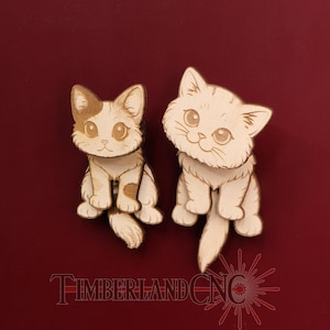 May include: Two light-colored wooden cat cutouts with engraved details, featuring large eyes and pointed ears. The left cat has a small spot on its head, while the right cat has a fluffy appearance. The text TIMBERLANDCNC is visible below.